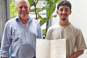 Zwei Männer stehen nebeneinander in einem Raum mit Glasdach, einer hält eine geöffnete Urkunde mit Text, im Hintergrund eine große grüne Pflanze.