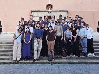Eine größere Gruppe von 29 Menschen steht in mehreren Reihen auf einer Außentreppe vor einem Gebäude mit rosa Fassade, das eine kleine runde Öffnung über der Tür und eine Hausnummer 4 zeigt.