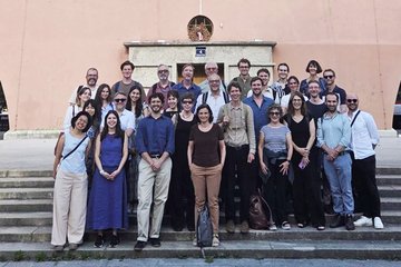 Eine größere Gruppe von 29 Menschen steht in mehreren Reihen auf einer Außentreppe vor einem Gebäude mit rosa Fassade, das eine kleine runde Öffnung über der Tür und eine Hausnummer 4 zeigt.