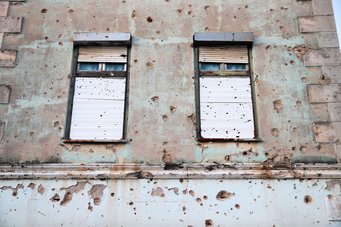 The exterior wall of a building with two windows, marked by numerous bullet holes and signs of gunfire; significant damage to the stucco and masonry is visible.