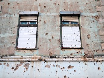 Außenwand eines Gebäudes mit zwei Fenstern, die von zahlreichen Einschusslöchern und Spuren von Beschuss geprägt ist, deutliche Schäden an Putz und Mauerwerk sichtbar.