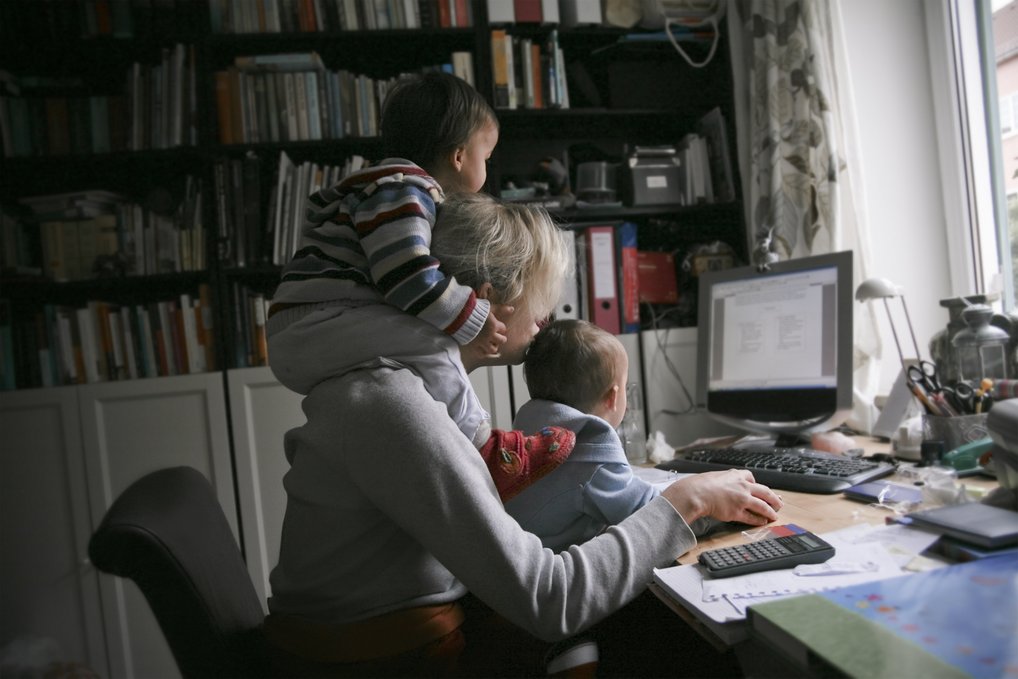 In einem Arbeitszimmer mit Büchern im Hintergrund sitzt eine Frau vor einem Computer, begleitet von zwei kleinen Kindern, die auf ihrem Schoß und ihrer Schulter sitzen.