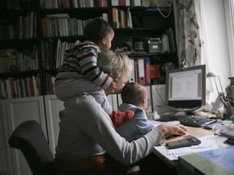 In einem Arbeitszimmer mit Büchern im Hintergrund sitzt eine Frau vor einem Computer, begleitet von zwei kleinen Kindern, die auf ihrem Schoß und ihrer Schulter sitzen.