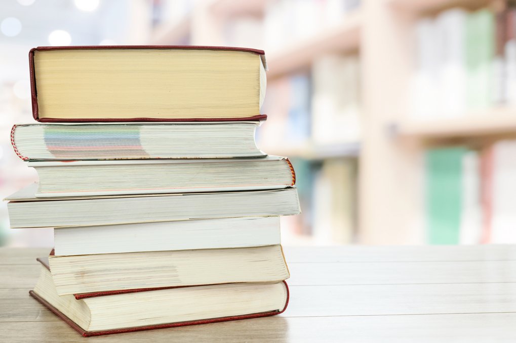 Mehrere dicke Bücher sind auf einem hellen Holztisch übereinander gestapelt, im Hintergrund verschwommene Bücherregale in einer Bibliothek.