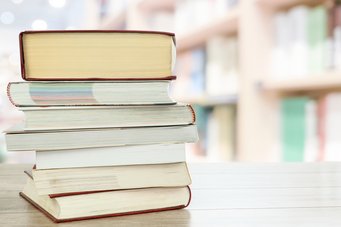 Several thick books are stacked on top of one another on a light-colored wooden table; in the background, bookshelves in a library are blurred.