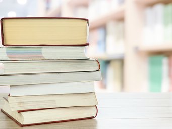 Mehrere dicke Bücher sind auf einem hellen Holztisch übereinander gestapelt, im Hintergrund verschwommene Bücherregale in einer Bibliothek.
