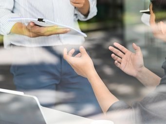 Zwei Personen führen eine Diskussion, eine steht mit Klemmbrett, die andere sitzt und verwendet Gesten zur Erklärung.