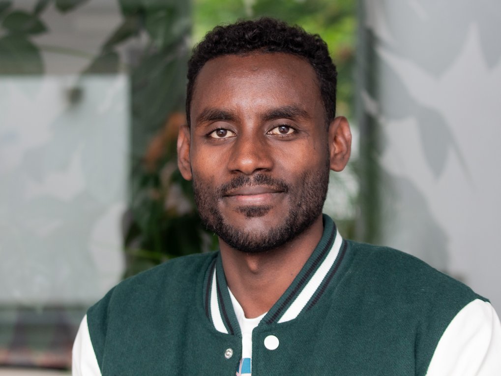 Portrait of a man with dark brown skin, short curly hair, and a beard, wearing a green college jacket with white sleeves, against a blurred background.