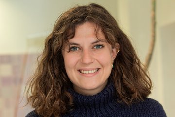A woman with shoulder-length, curly brown hair is wearing a dark blue knit sweater and smiling directly at the camera; the background is out of focus.