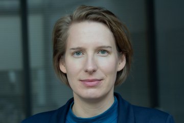 Auf dem Foto ist ein dem Anschein nach feminines Exemplar der Gattung Homo sapiens intellectualis zu erkennen. Es trägt das Haar kurz, die Ohren sind zum größten Teil freigelegt, das dunkelblaue Sakko und der im Blauton etwas heller gehaltene Pullover lassen den Hals offen. Das Individuum ist sitzend oder stehend vor einem Vorhang platziert. Die Szene ist dunkel gehalten.