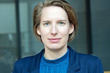 Porträt einer Frau mit kurzem Haar und blauem Oberteil vor unscharfem Hintergrund.