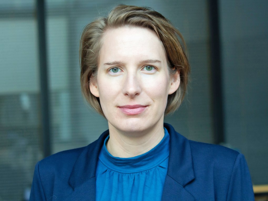 Porträt einer Frau mit kurzem Haar und blauem Oberteil vor unscharfem Hintergrund.