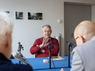 Älterer Mann in rotem Pullover, Wolfgang Streeck, spricht bei einem Interview mit zwei weiteren Personen.