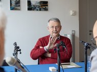 Wolfgang Streeck in red sweater speaking at a table with microphones.