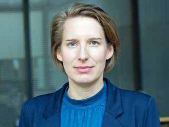 Professional portrait of an individual with short light brown hair, green eyes, and neutral expression, wearing a blue blazer and blouse, with an indistinct indoor background.