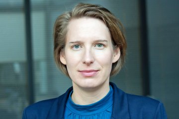 Professional portrait of an individual with short light brown hair, green eyes, and neutral expression, wearing a blue blazer and blouse, with an indistinct indoor background.