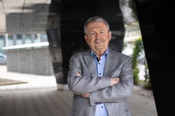 Prof. em. Dr. Dr. h.c. Wolfgang Streeck Älterer Mann mit grauem Sakko und blauem Hemd posiert mit verschränkten Armen vor einem modernen, urbanen Gebäude mit dunklen und hellen Flächen.