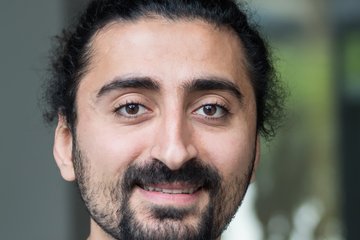 Eine Person hat dunkles lockiges Haar und trägt ein hellblaues Hemd. Der Hintergrund ist unscharf, was auf eine Innenraumumgebung mit sanfter Beleuchtung hindeutet.