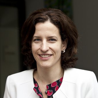 Cornelia Woll A woman dressed in a white blazer and a red-black patterned blouse, featuring a pearl pendant necklace and small hoop earrings, stands indoors.