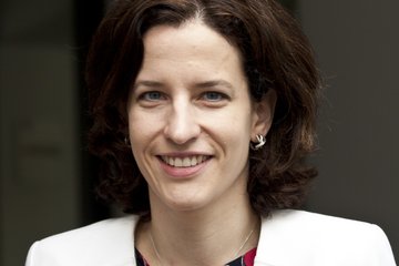 Eine Frau in einem weißen Blazer und einer rot-schwarz gemusterten Bluse, mit einer Perlenkette und kleinen Creolen, steht in einem Innenraum.