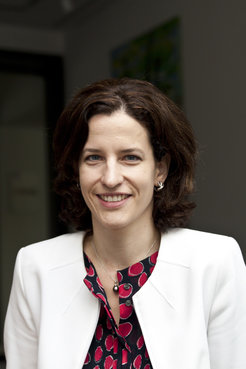 A woman dressed in a white blazer and a red-black patterned blouse, featuring a pearl pendant necklace and small hoop earrings, stands indoors.