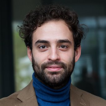 Donato Di Carlo Die Person hat lockiges Haar, trägt einen blauen Rollkragenpullover und einen braunen Blazer mit einem gemusterten Einstecktuch; der Hintergrund ist unscharf.