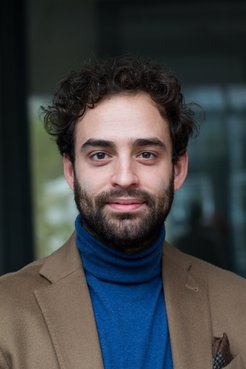 Die Person hat lockiges Haar, trägt einen blauen Rollkragenpullover und einen braunen Blazer mit einem gemusterten Einstecktuch; der Hintergrund ist unscharf.