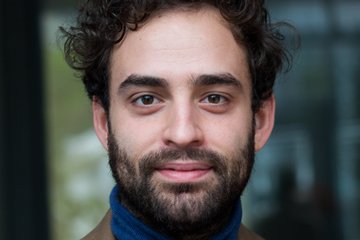 Die Person hat lockiges Haar, trägt einen blauen Rollkragenpullover und einen braunen Blazer mit einem gemusterten Einstecktuch; der Hintergrund ist unscharf.