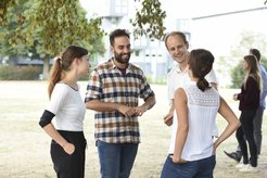 A group of four people are standing in a park, talking, surrounded by several bicycles and trees in the background.