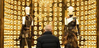 Glänzende Aussicht: Wer diese Kleidung trägt, rückt ins Rampenlicht, suggeriert das Schaufenster. So hebt die Modefirma hervor, dass ihre Produkte als Statussymbol dienen. Zwei Personen stehen vor einem Schaufenster mit leuchtenden, goldenen Lichtern, in dem zwei Schaufensterpuppen in modischer, schwarzer Kleidung präsentiert werden.