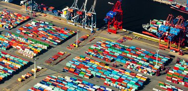 Luftaufnahme eines geschäftigen Containerhafens mit farbigen Containern, großen Kränen und Schiffen, die im Hintergrund beladen werden.