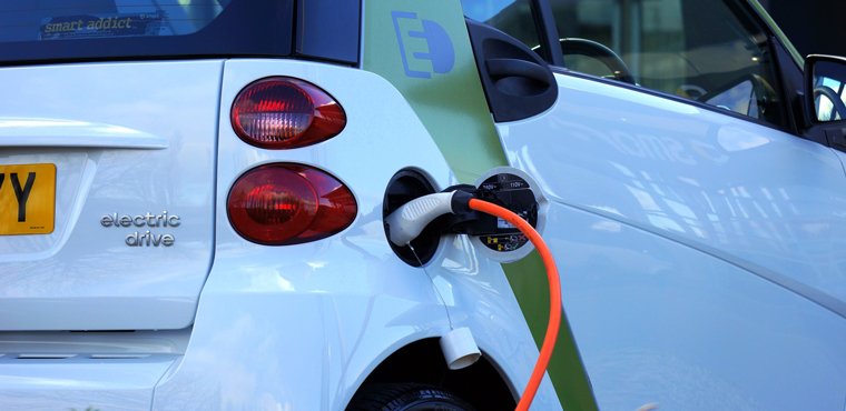 Zwischen Offenheit und Festlegung: Ein Dilemma in der Förderung von Innovationen Rückansicht eines Elektroautos mit angeschlossenem Ladekabel, das an einer Ladestation aufgeladen wird. Das Fahrzeug hat weiße Karosserie und grüne Akzente.