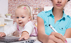 Frau mit türkisfarbener Bluse schreibt mit Stift, während das Kind neben ihr auf die Laptop-Tastatur zeigt. Im Hintergrund eine Backsteinwand und weiße Küchenelemente.