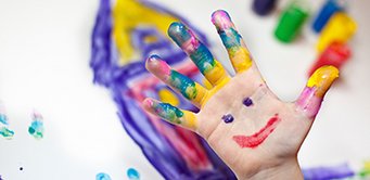 Eine Kinderhand ist in verschiedenen Farben bemalt, mit einem lächelnden Gesicht auf der Handfläche. Im Hintergrund sind unscharf Malutensilien und ein gemaltes Haus zu sehen.