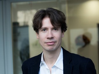 Leon Wansleben, Leiter der „Forschungsgruppe Soziologie öffentlicher Finanzen und Schulden“ am MPIfG Eine Person trägt einen dunklen Anzug und ein weißes Hemd mit offenem Kragen, steht vor einem modernen Gebäude mit Glasfront.