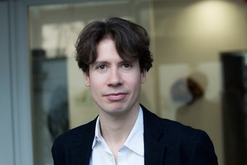 Eine Person trägt einen dunklen Anzug und ein weißes Hemd mit offenem Kragen, steht vor einem modernen Gebäude mit Glasfront.