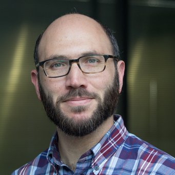 Nahaufnahme eines Mannes mit Bart, Brille und kariertem Hemd, der vor einem unscharfen Hintergrund lächelt.
