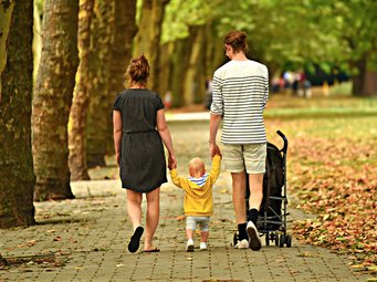 Ein Paar läuft mit einem Kleinkind durch einen herbstlichen Park. Das Kind trägt eine gelbe Jacke und wird von beiden Eltern an der Hand gehalten. Ein Kinderwagen wird nebenhergeschoben.