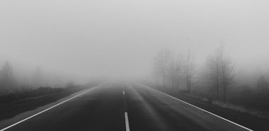 Eine gerade Straße zieht sich in einen dichten Nebel hinein, während kahle Bäume am Straßenrand im Hintergrund leicht sichtbar sind.