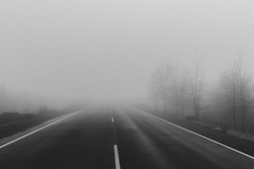Eine gerade Straße zieht sich in einen dichten Nebel hinein, während kahle Bäume am Straßenrand im Hintergrund leicht sichtbar sind.