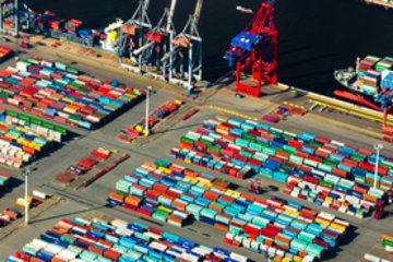 Luftaufnahme eines geschäftigen Containerhafens mit farbigen Containern, großen Kränen und Schiffen, die im Hintergrund beladen werden.