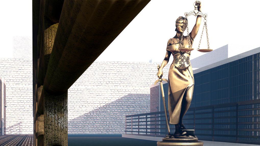 Goldene Statue der Justitia, die eine Waage in der rechten und ein Schwert in der linken Hand hält, steht in einem modern urbanen Raum, umgeben von hohen Gebäuden und einer Steinmauer.