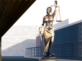 Goldene Statue der Justitia, die eine Waage in der rechten und ein Schwert in der linken Hand hält, steht in einem modern urbanen Raum, umgeben von hohen Gebäuden und einer Steinmauer.