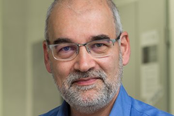 Ein Mann mit Bart und Brille, der ein blaues Hemd trägt, lächelt in die Kamera.