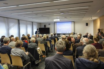 Die Teilnehmer sitzen in Reihen in einem modernen Konferenzraum und konzentrieren sich auf einen Redner, der ein Thema präsentiert, das auf einer großen Leinwand angezeigt wird.