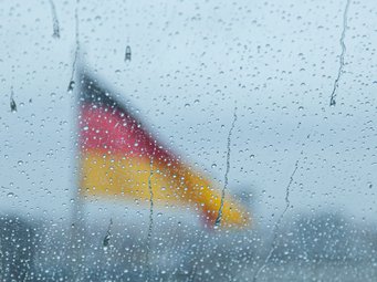 Durch eine mit Regentropfen bedeckte Scheibe ist eine unscharf erscheinende Deutschlandfahne zu erkennen, die im Hintergrund weht.