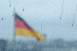 Durch eine mit Regentropfen bedeckte Scheibe ist eine unscharf erscheinende Deutschlandfahne zu erkennen, die im Hintergrund weht.