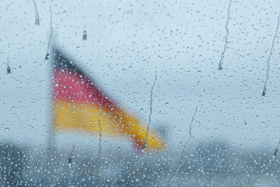 Durch eine mit Regentropfen bedeckte Scheibe ist eine unscharf erscheinende Deutschlandfahne zu erkennen, die im Hintergrund weht.