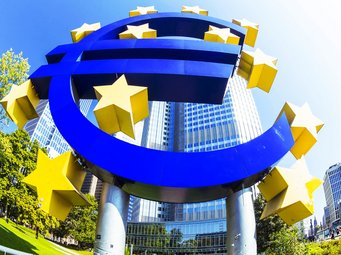 Großes blaues Euro-Symbol, umgeben von gelben Sternen, steht vor einer Kulisse aus modernen Hochhäusern und grünen Bäumen bei klarem Himmel.
Mann mit lockigem, braunem Haar träg
