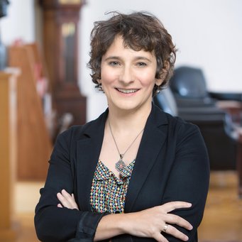 Marion Fourcade Auf dem Bild ist eIn vermutlich weibliches Exemplar der Gattung Homo sapiens in einem leicht verschwommen aufgenommenen Raum zu erkennen. Es trägt das dunkelblonde halblange Haar offen, lächelt freundlich und empathisch dem Betrachter entgegen, dabei die Arme verschränkt unterhalb der Brust gehalten. Die dunkle Jacke lässt unterhalb des Halses genügend Raum für den vollständig sichtbaren Kettenschmuck. Links im Bilde befinden sich auf Podesten mindestens zwei Büsten, die näher zu beschreiben nicht möglich ist.